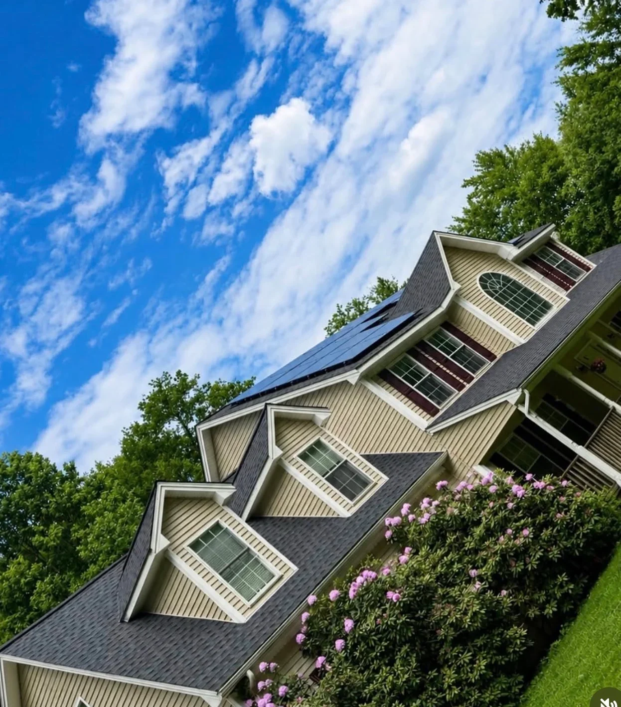 Solar panels installed on dark gray shingled roof with white dormers and purple flowering shrubs