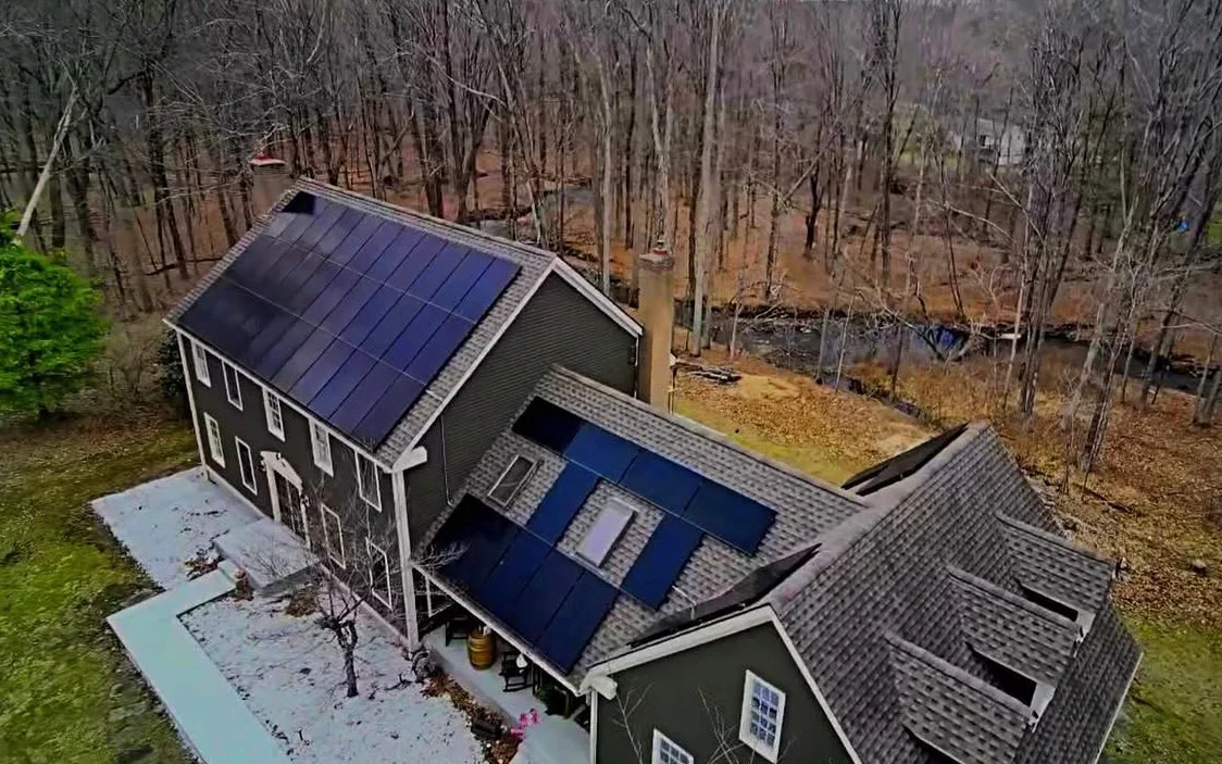 Aerial view of home with dark blue solar panels installed on multiple roof sections in wooded setting